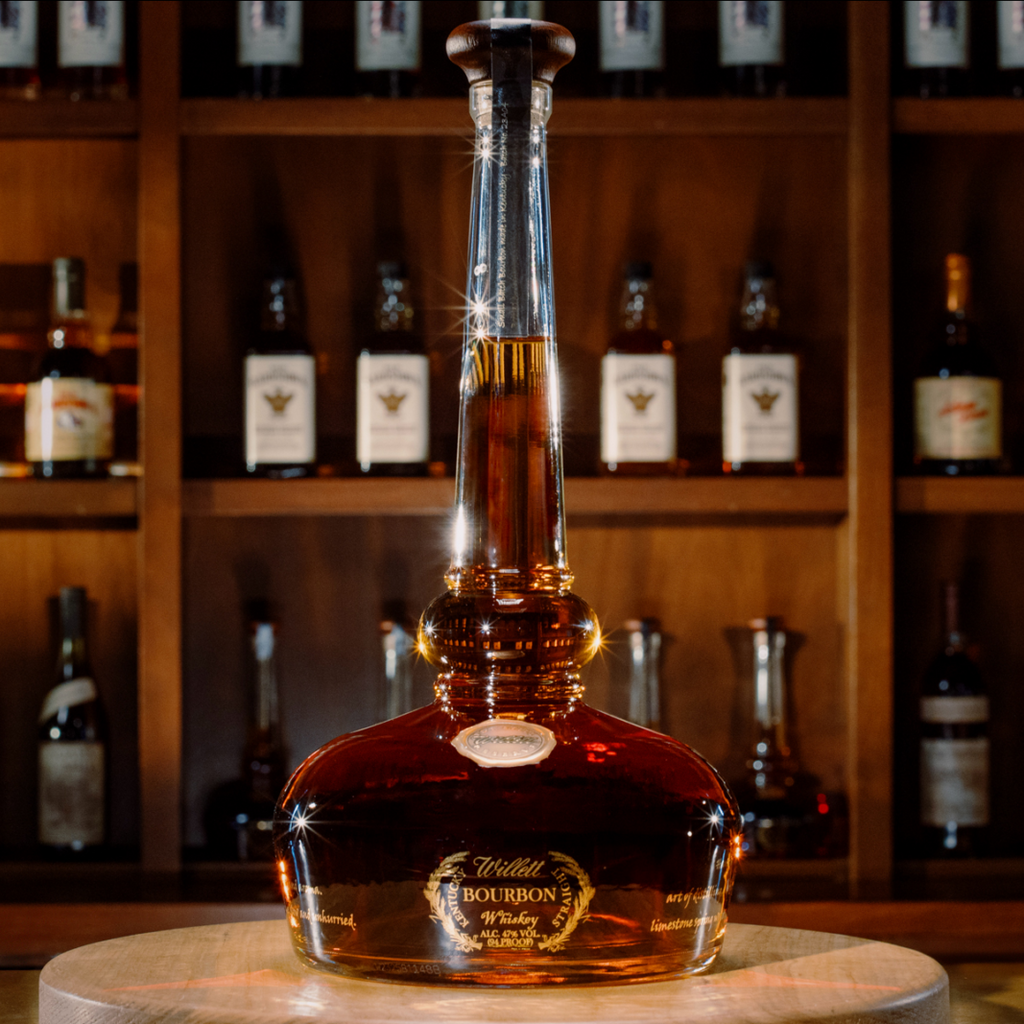 Bottle of Willet Bourbon on a wooden stand with shelves of bottles in the background